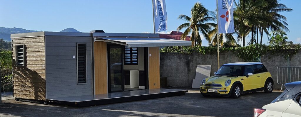 Photo d'un conteneur habillé en bardage fibre-ciment, transformé en espace modulaire moderne avec des volets de ventilation et un store. L'installation est accompagnée d'une voiture Mini Cooper jaune, illustrant l'échelle et l'intégration du conteneur dans un environnement extérieur tropical.